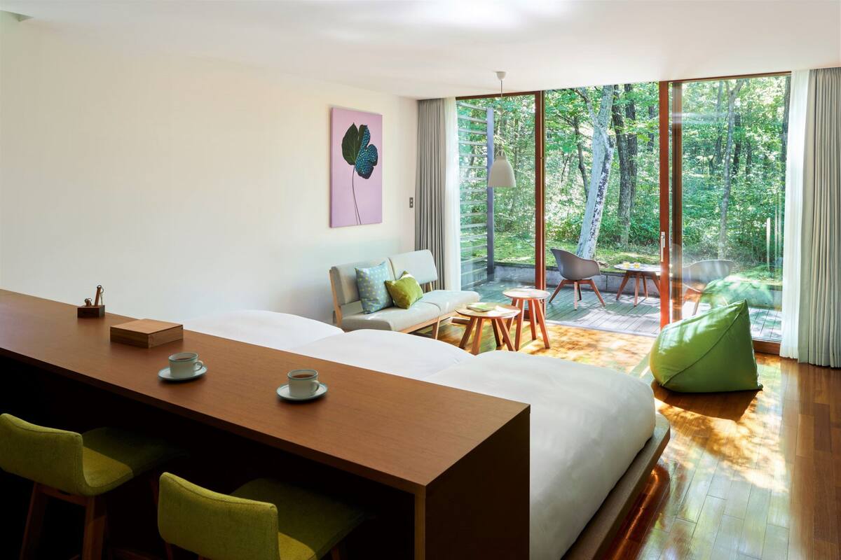 A modern room with a wooden desk, green chairs, a sofa, and a view of a forest through large windows.