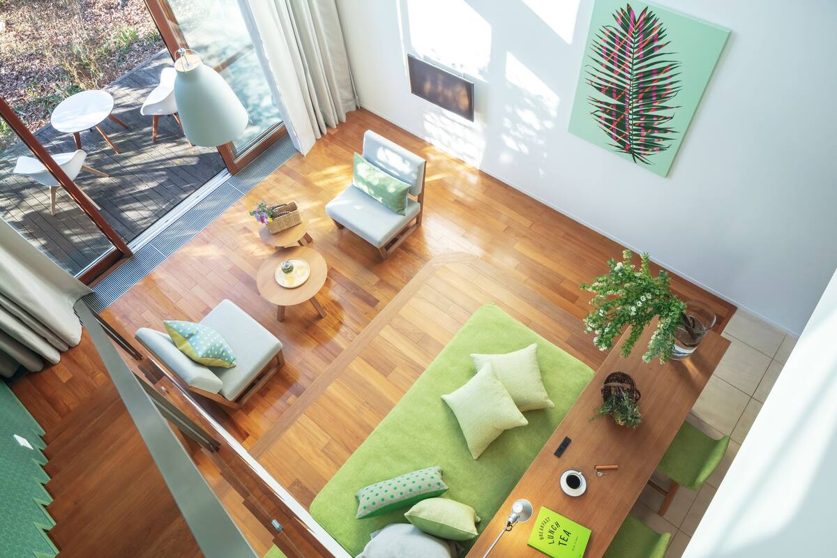 A modern living room with a wooden floor, a green sofa, a small table with a plant, and a view of the outdoors through large windows.