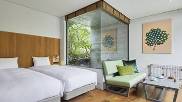A modern hotel room with two beds, a glass wall with a view of greenery, a small table with a mirror, and a pet bed.