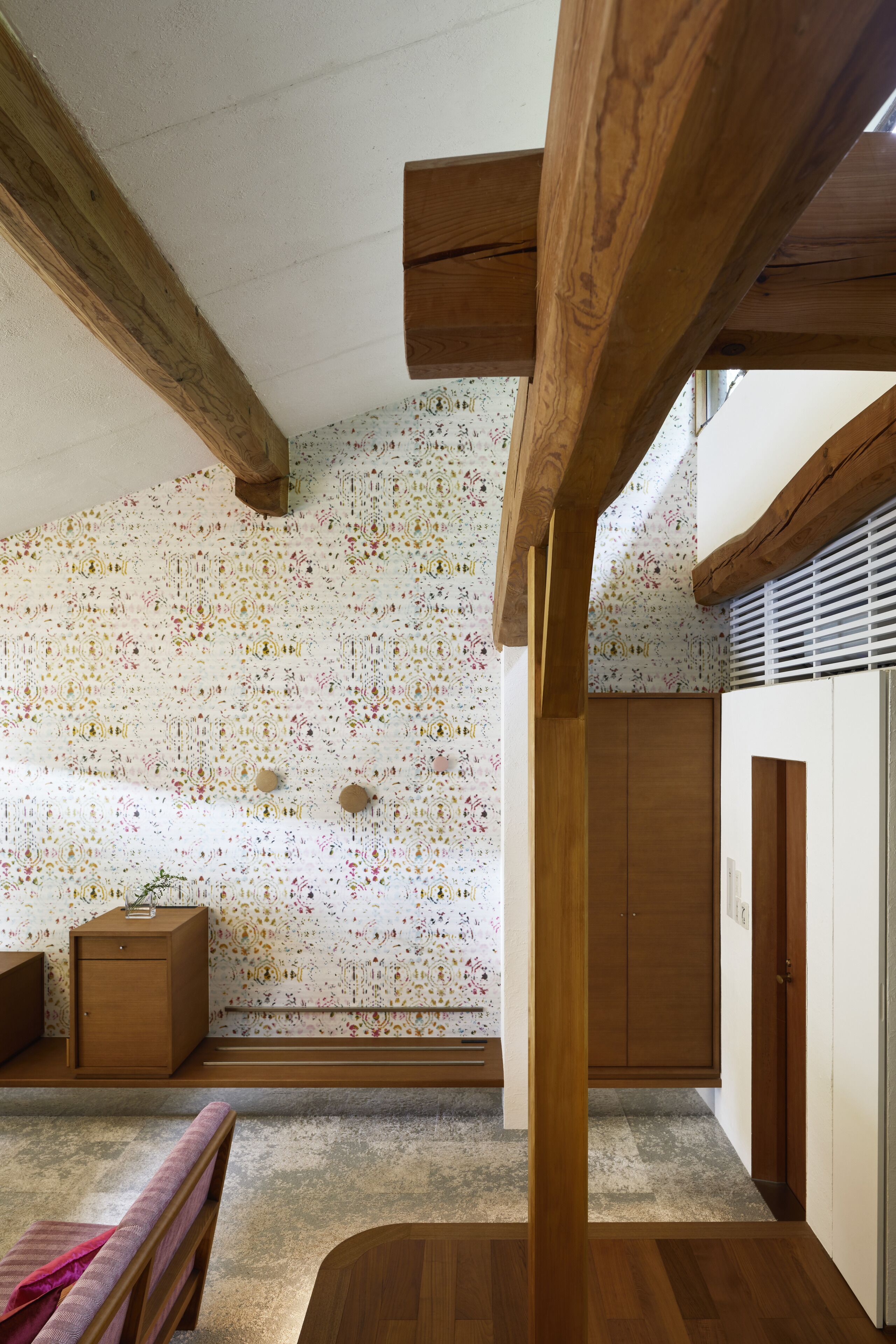 A room with wooden beams, a patterned wall, a bed, a wardrobe, and a window with blinds.