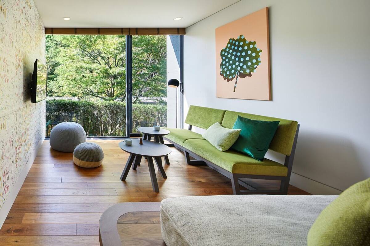 A modern living room with a green sofa, round ottomans, a coffee table, and a large window offering a view of greenery.
