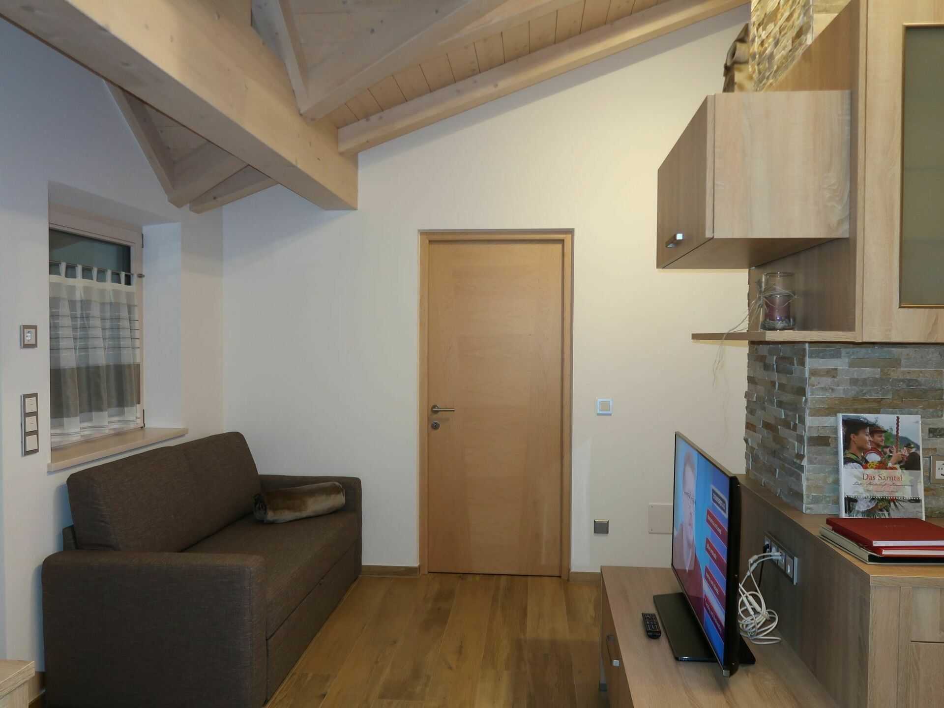 Una habitación acogedora con techo de madera, un sofá marrón, una puerta de madera y un televisor sobre un mueble de madera.
