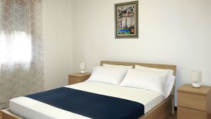 A neatly made bed with a blue blanket, two white pillows, and a wooden headboard. There are two bedside tables with lamps, and a framed picture hangs on the wall above the bed. - Nice apartment for 6 guests with WIFI, TV, patio and pets allowed (Eraclea)