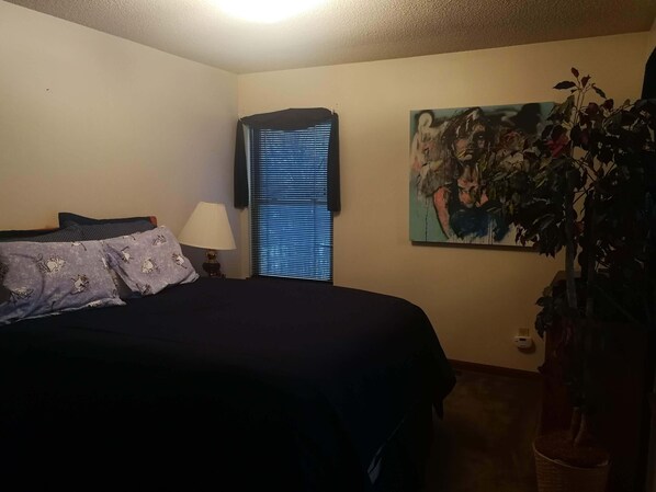 Une chambre à coucher avec un lit, des oreillers, une lampe, une fenêtre avec des stores, un tableau au mur et une plante en pot.