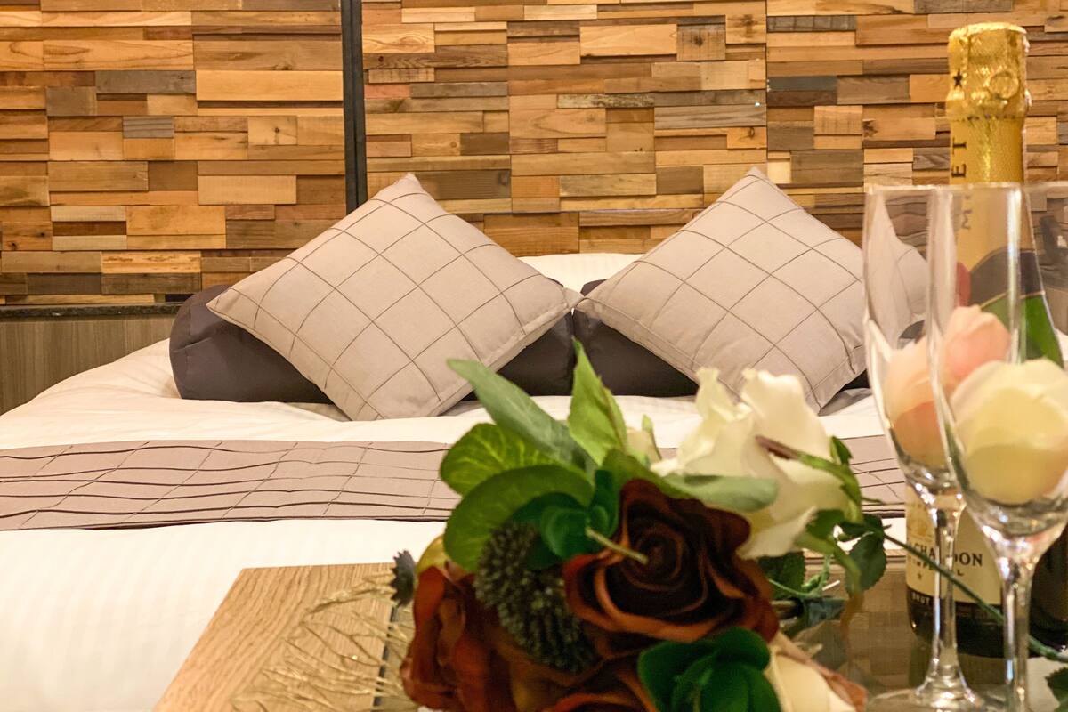 A neatly made bed with two pillows, a vase with flowers, and two wine glasses on a table.