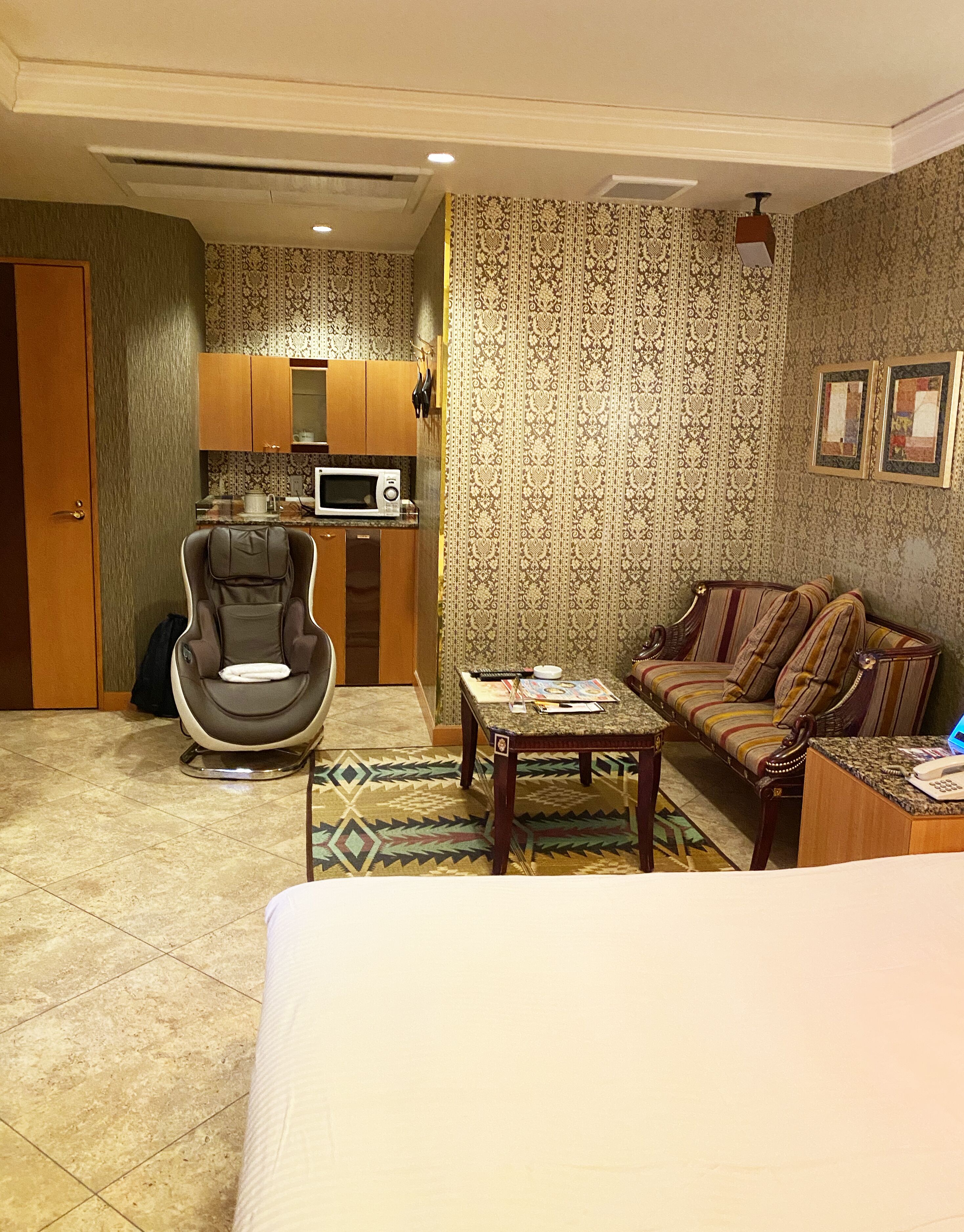 A hotel room with a bed, a massage chair, a small table, a sofa, a microwave, and a kitchen area.