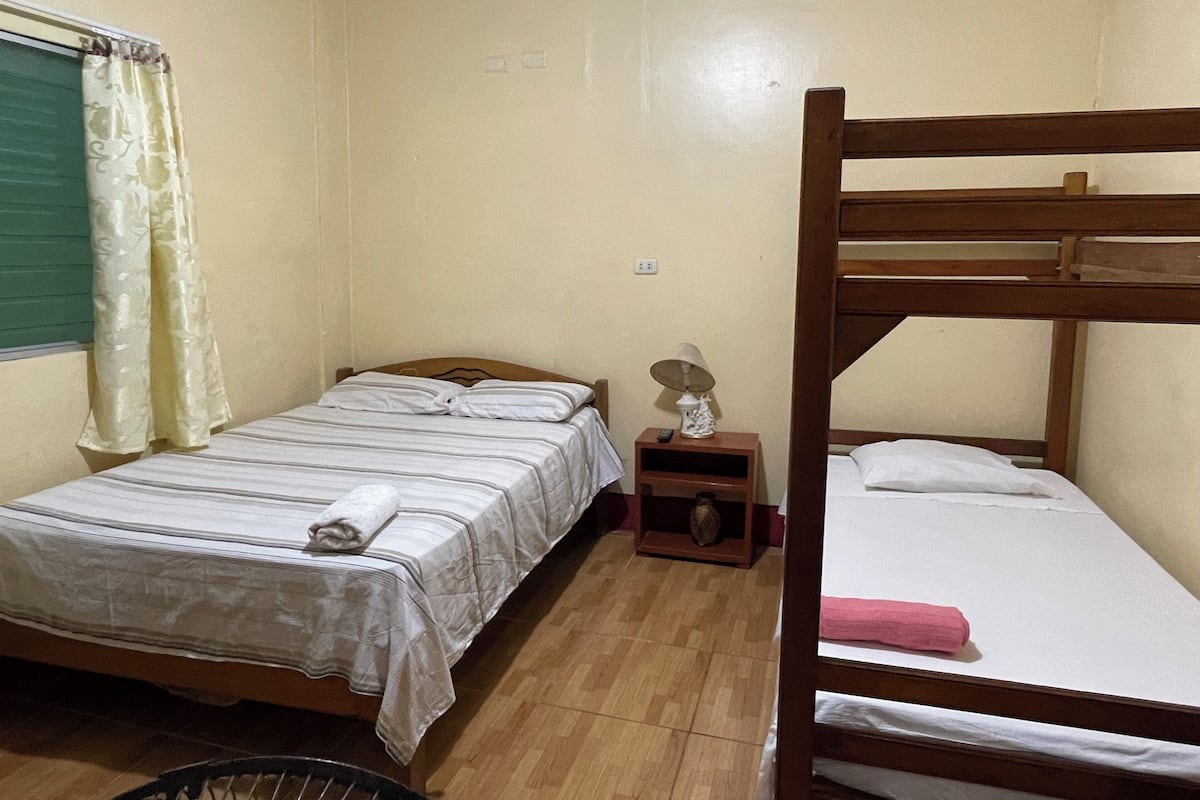 Un alojamiento pequeño de una sola habitación con dos camas, una litera de madera, una mesita de noche con lámpara y una ventana con cortinas.