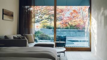 A modern hotel room with a large sliding glass door leading to a balcony, a bed, a seating area, and a view of autumn foliage.