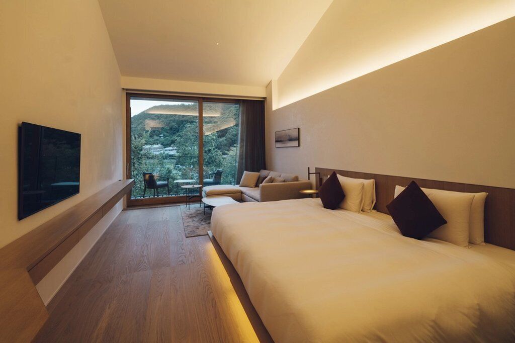 A modern hotel room with a large bed, a sofa, a TV, and a view of a mountainous landscape through the window.