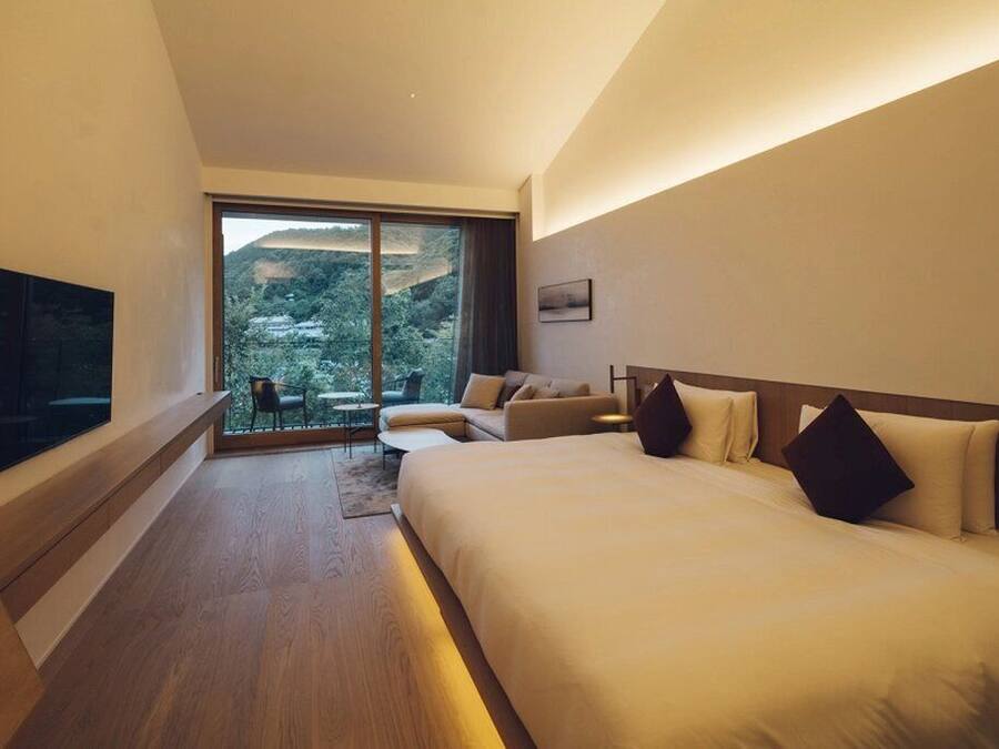 Quarto de hotel moderno com uma cama grande, um sofá, uma TV e vista para uma paisagem montanhosa pela janela.