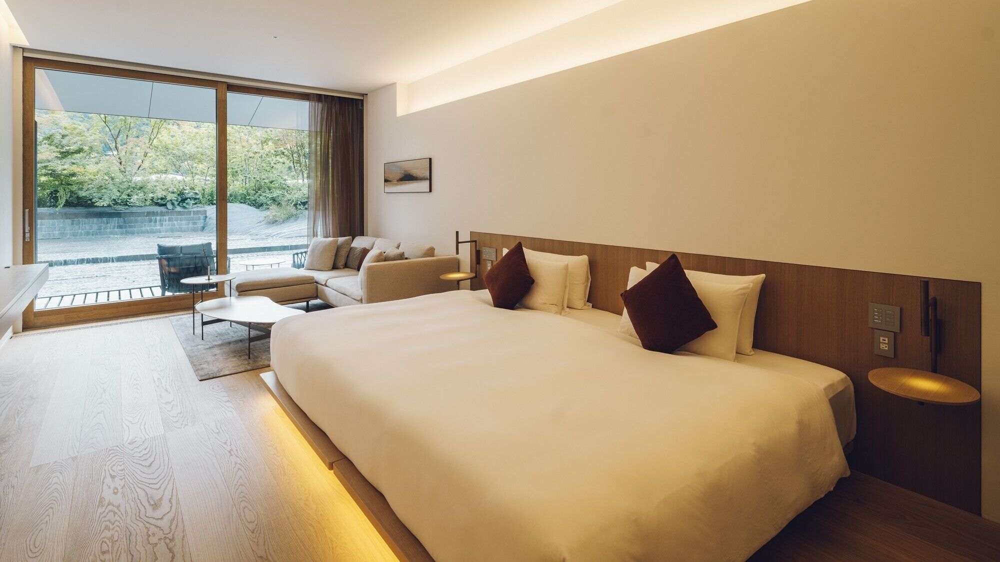 A modern hotel room with a large bed, a sofa, a small table, and a view of greenery outside.