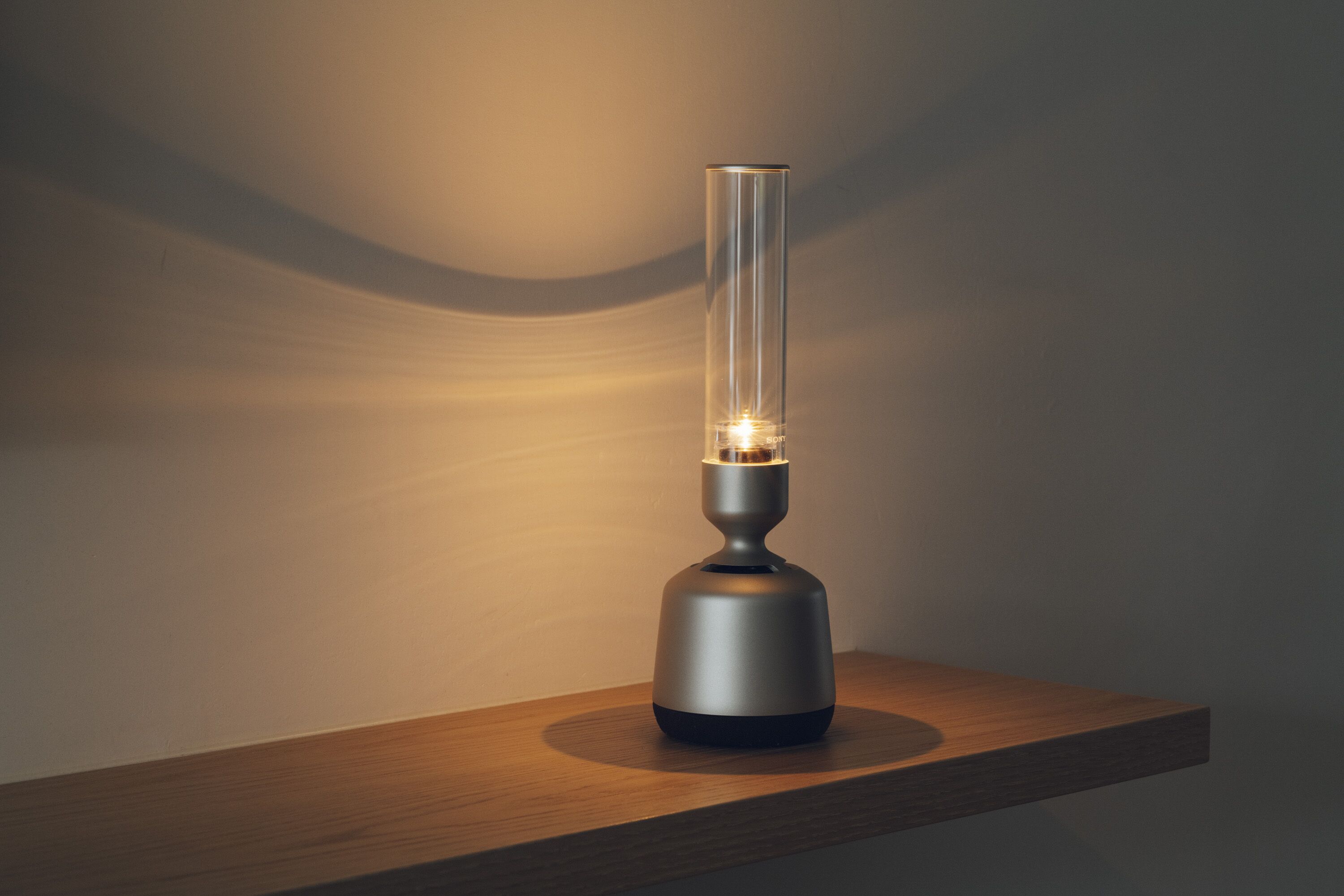 A modern, minimalist table lamp with a cylindrical glass shade and metallic base, placed on a wooden surface against a plain wall.