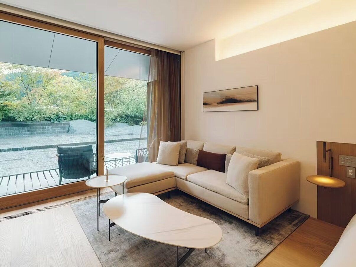 A modern hotel room with a large window, a sofa, a small table, and a wooden floor.