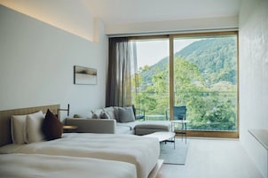 A modern hotel room with a large bed, a sofa, a small table, and a balcony with a view of a forested mountain.