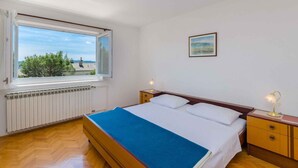 Een slaapkamer met een groot raam, een bed met een blauwe deken, een nachtkastje met een lamp en een lijstje met een foto aan de muur.