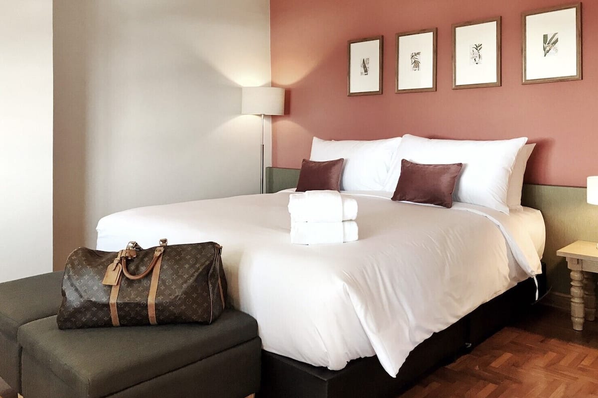 Hotellihuone, jossa on suuri sänky, yöpöytä, lamppu, tuoli ja Louis Vuitton -merkkinen laukku.