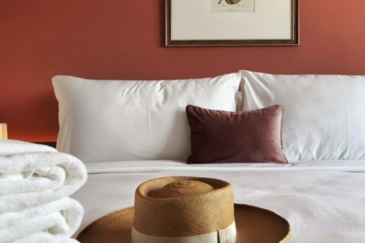 A bed with white bedding, a brown pillow, and a straw hat on a round table.