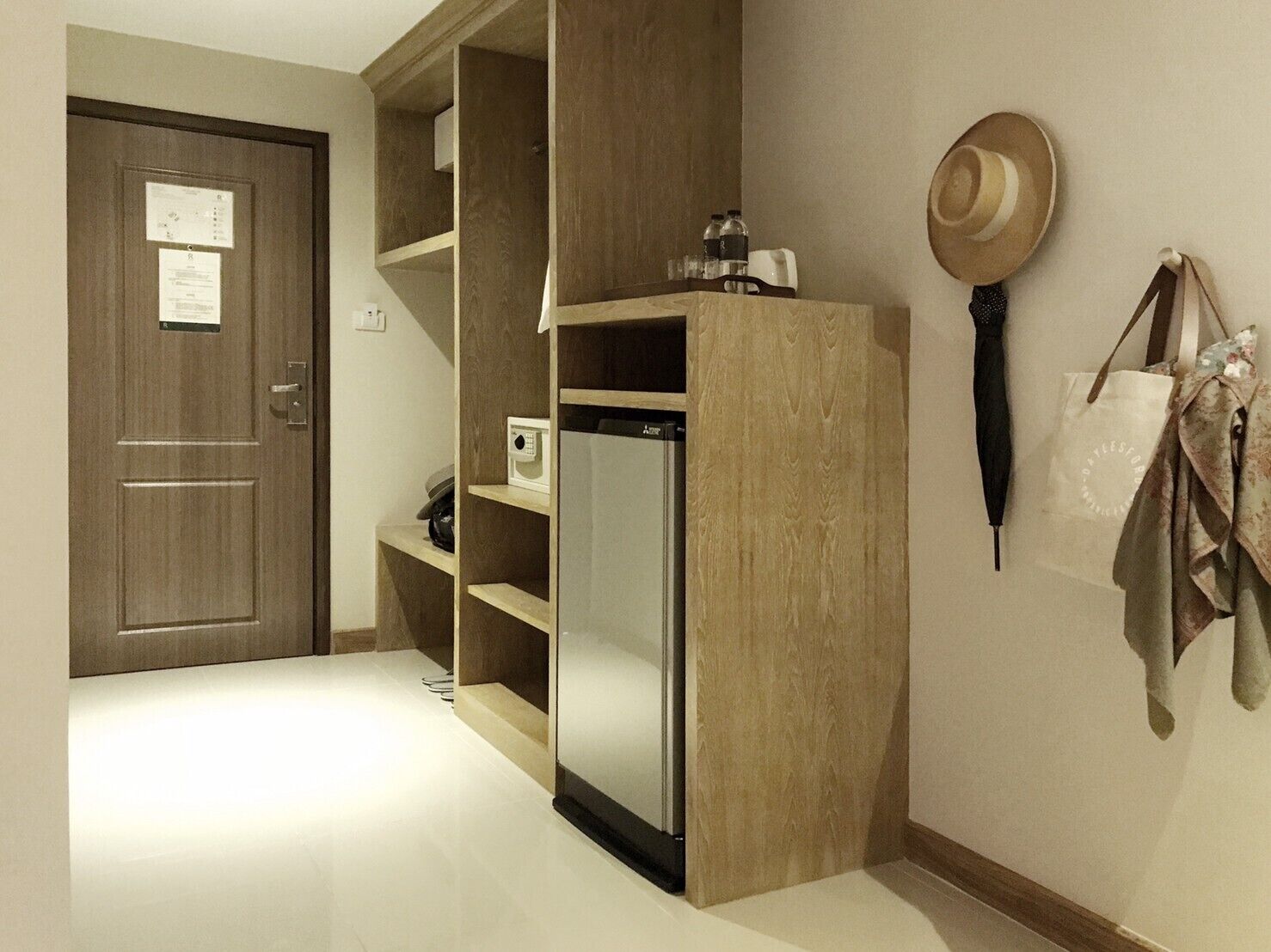 A hotel room with a wooden wardrobe, a microwave, a refrigerator, a door with a notice, and a hat on the wall.