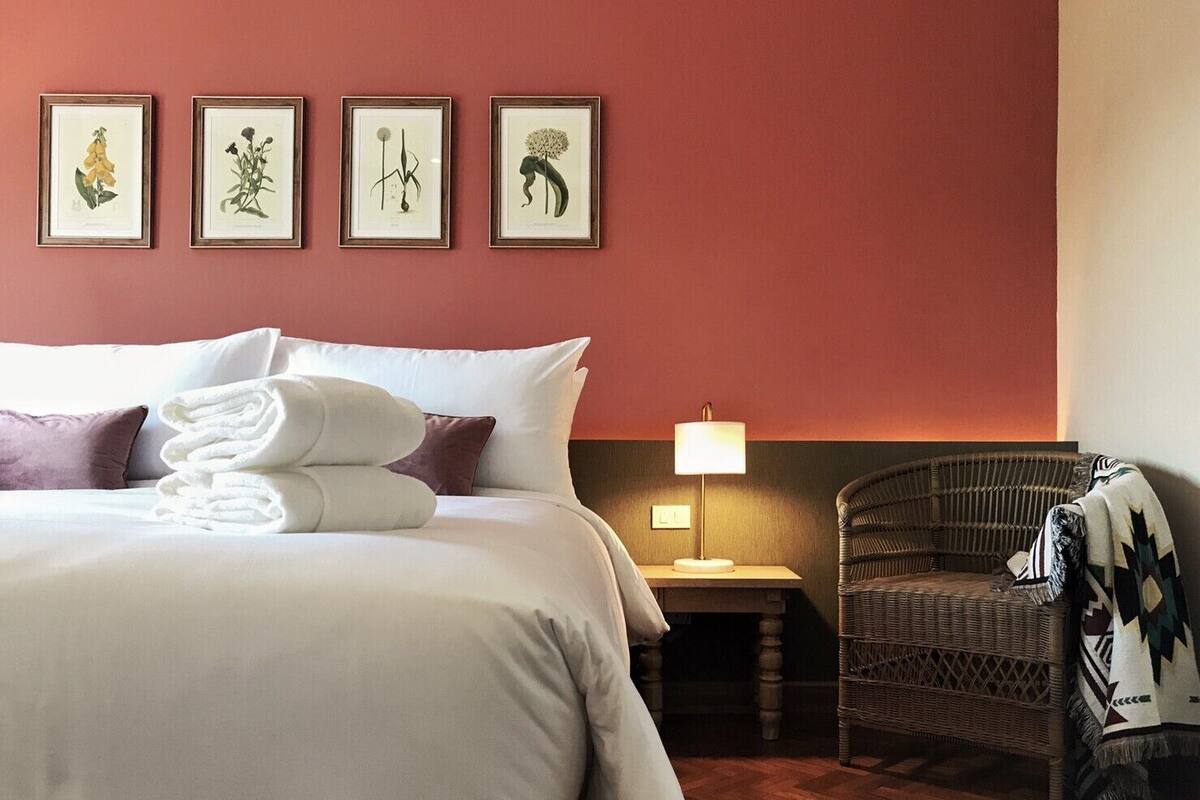 Hotellihuone, jossa on suuri sänky, yöpöytä lampun kanssa, rottinkituoli ja seinällä neljä kehystettyä kasvitieteellistä taideteosta.