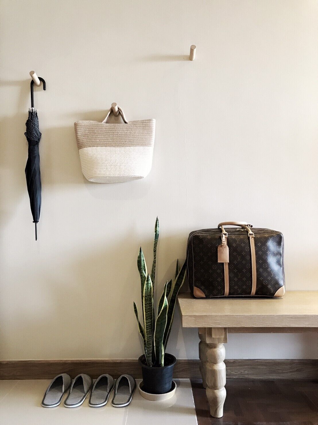 A wall-mounted umbrella, a woven basket, a potted plant, a Louis Vuitton bag, and a pair of slippers on a wooden bench.