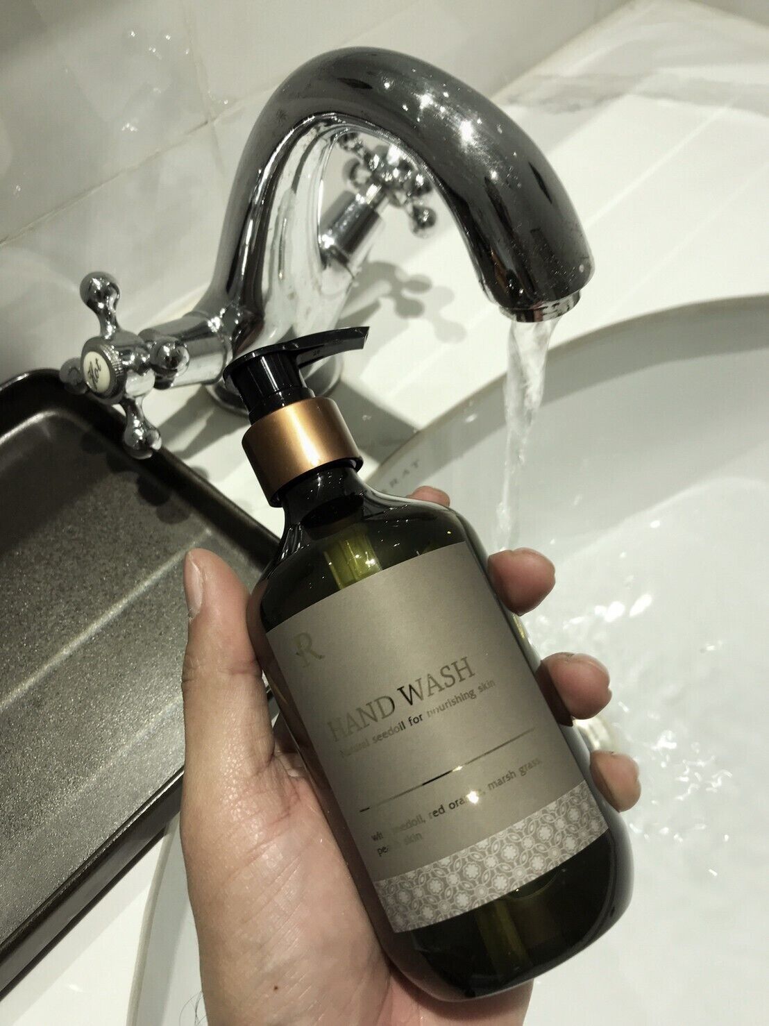 A hand holding a bottle of hand wash soap under a running faucet in a bathroom.