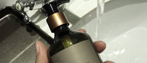 A hand holding a bottle of hand wash soap under a running faucet in a bathroom.
