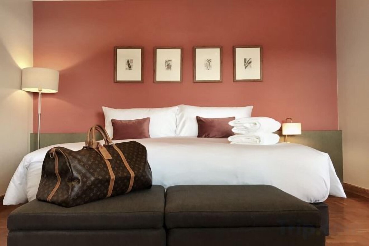 A hotel room with a bed, pillows, a bedside lamp, a lamp on the floor, a large bag, and a wall with framed pictures.