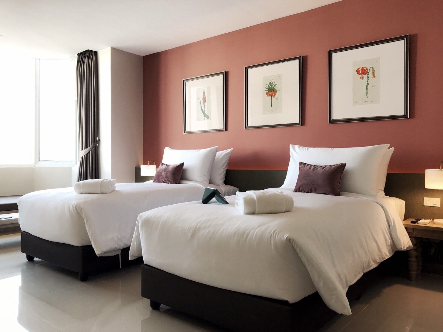 A hotel room with two beds, a large window with curtains, and three framed botanical prints on the wall.