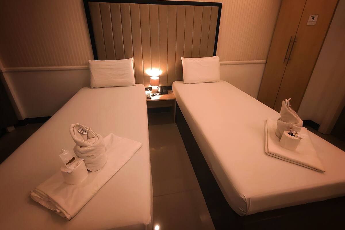 Quarto de hotel com duas camas de solteiro, cada uma com travesseiro, luminária de cabeceira e toalha dobrada na cama.