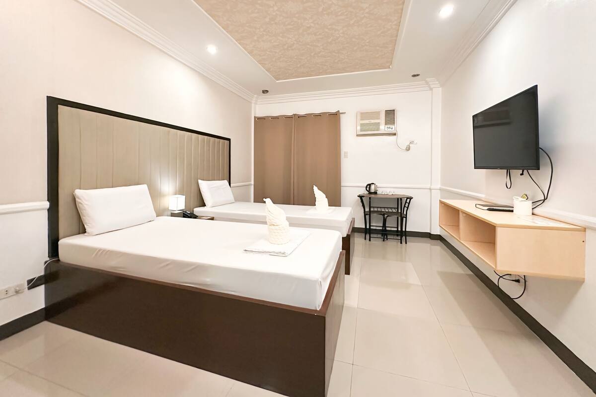 Quarto de hotel com duas camas, uma escrivaninha, uma televisão e um ventilador de teto.