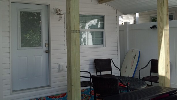 Un patio techado con una pérgola de madera, una mesa de comedor con sillas, una tabla de surf y una alfombra colorida.