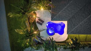 Una piscina con interior azul, rodeada de palmeras y una zona pavimentada con dos bancos.