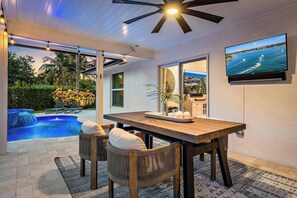 A poolside dining area with a table and chairs, a ceiling fan, and a TV mounted on the wall.