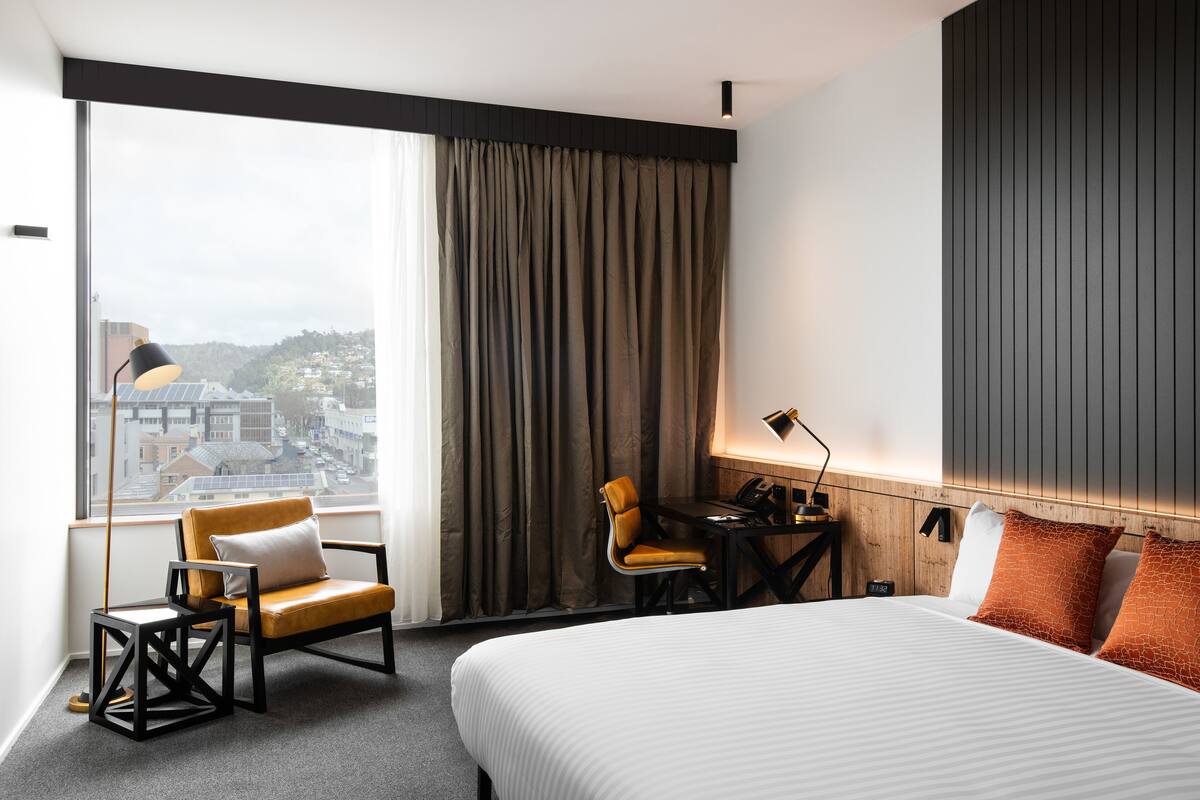 A hotel room with a large bed, a desk, a chair, and a view of the city through a large window.
