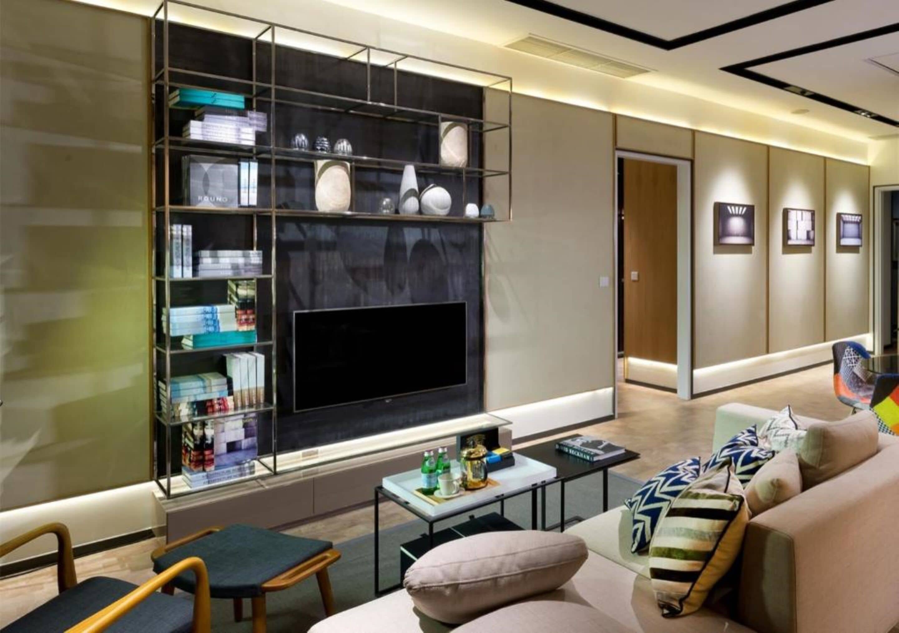 A modern hotel lobby with a large flat-screen TV, a glass display case with various items, a sofa with cushions, and a coffee table with drinks.