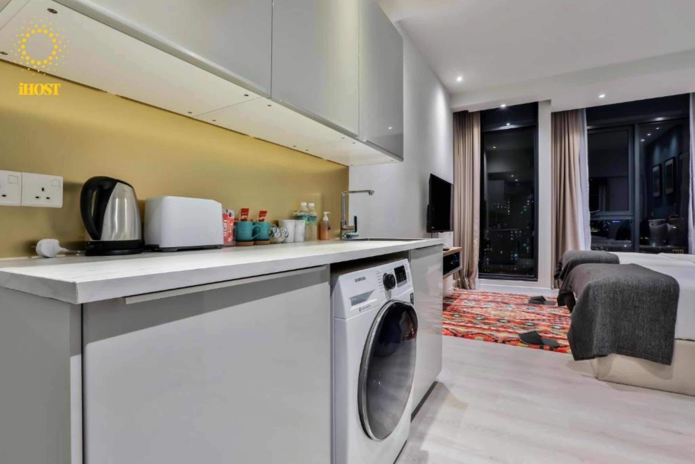 Ein modernes Hotelzimmer mit Waschmaschine, einer kleinen Küchenzeile, einem Bett und Blick über die Stadt durch das Fenster.