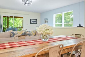 Un comedor con una mesa de madera, sillas y un jarrón de flores. Se ve un sofá con cojines estampados al fondo.