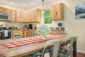 A kitchen with wooden cabinets, a stainless steel microwave, a dining table with a red and white checkered table runner, and a vase with white flowers.