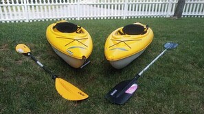 Two yellow kayaks with paddles and a paddleboard on a grassy lawn.