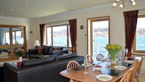 Sala de estar com sofá de couro preto, mesa de jantar posta para quatro pessoas e vista para o mar através de grandes janelas.