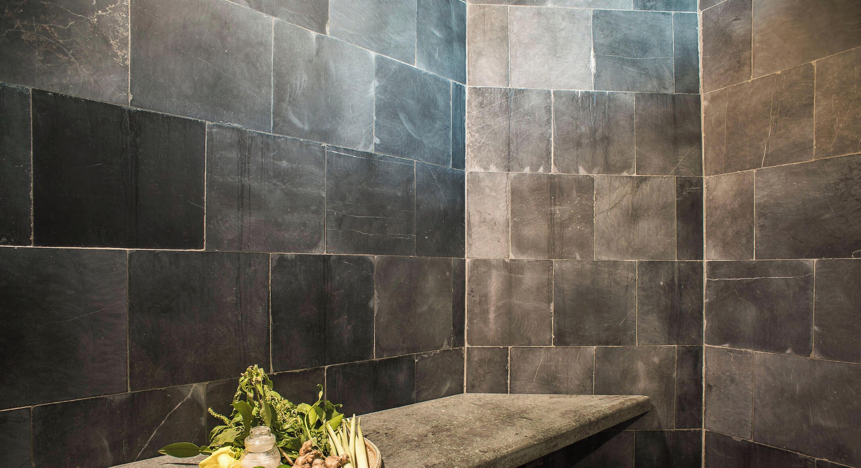 A bathroom with dark stone tiles and a bench with a tray of fresh herbs and lemons.