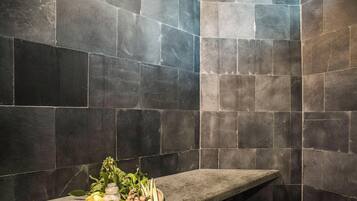 A bathroom with dark stone tiles and a bench with a tray of fresh herbs and lemons.