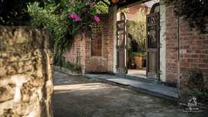 A brick building with a covered entrance, a stone wall, and flowering vines. - Le Clos Du Fil (Nam Hoa Lu)