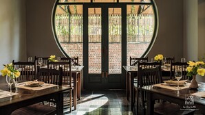 A dining area with a brick wall and a large circular window. Tables are set with plates, cutlery, and glasses. - Le Clos Du Fil (Nam Hoa Lu)
