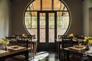 Comedor con pared de ladrillo y un amplio ventanal circular. Mesas dispuestas con platos, cubiertos y vasos.