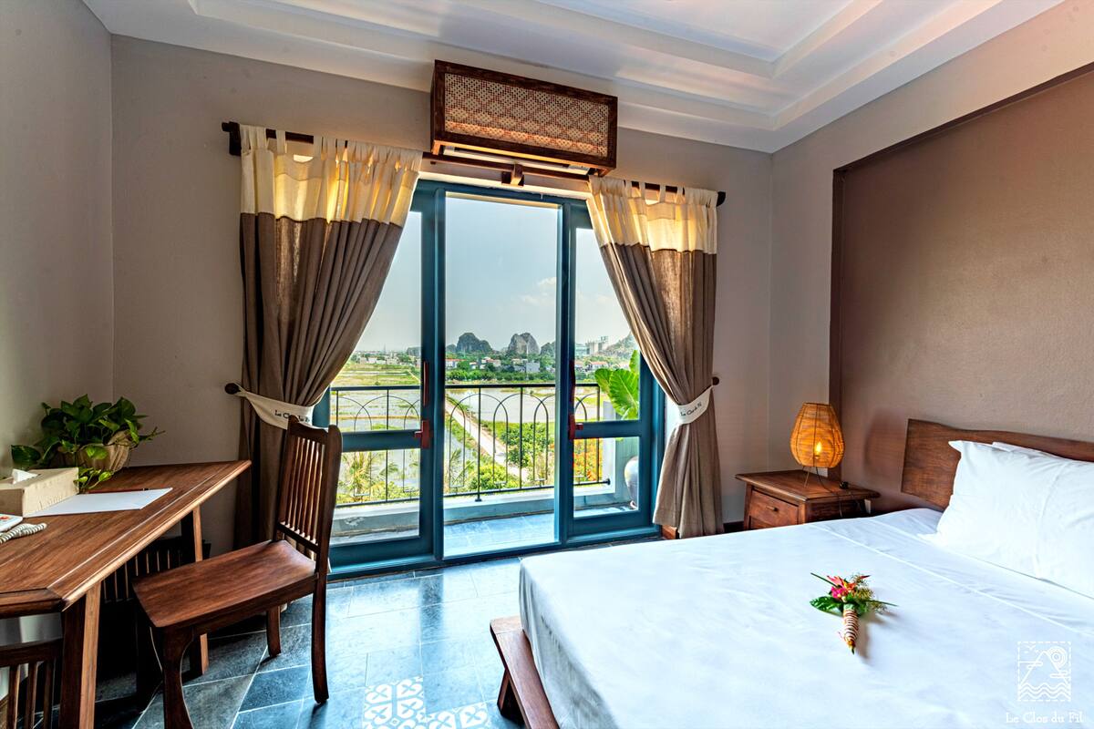 A hotel room with a bed, a desk with a chair, a nightstand with a lamp, and a view of the city through large windows.