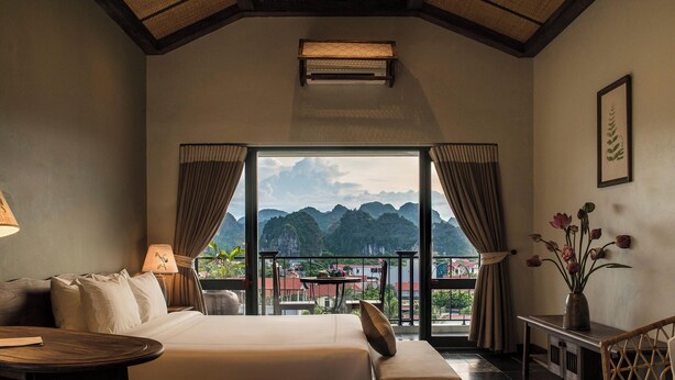 A bedroom with a large bed, a desk, a chair, and a view of mountains through a large window. - Le Clos Du Fil (Nam Hoa Lu)