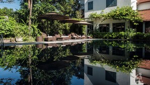 A serene pool area with lounge chairs and umbrellas, surrounded by lush greenery and a white building with a tiled roof. - Le Clos Du Fil (Nam Hoa Lu)