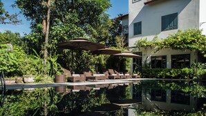 A serene pool area with lounge chairs and umbrellas, surrounded by lush greenery and a white building with a tiled roof. - Le Clos Du Fil (Nam Hoa Lu)