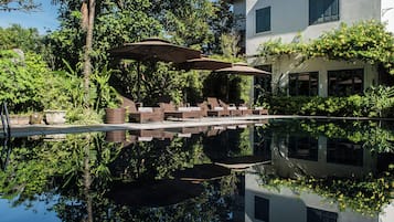 Uma área de piscina tranquila com espreguiçadeiras e guarda-sóis, rodeada por vegetação exuberante e um edifício branco com telhado em azulejos.